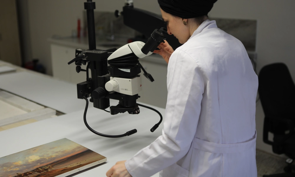 Person in a lab coat looking through the lens of a digital microscope into a painting