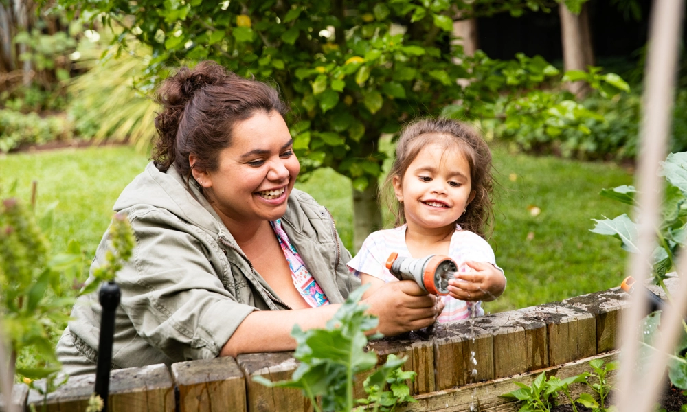 Woman and girl laughing while spraying water to plants in a garden bed