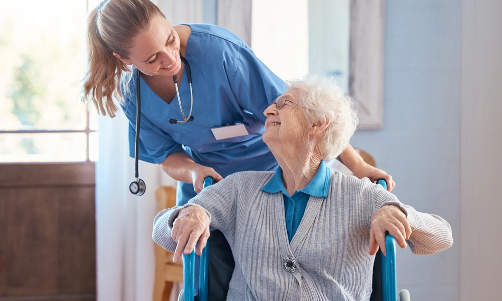 Person with a stethoscope around her neck is pushing an elderly person in a wheelchair